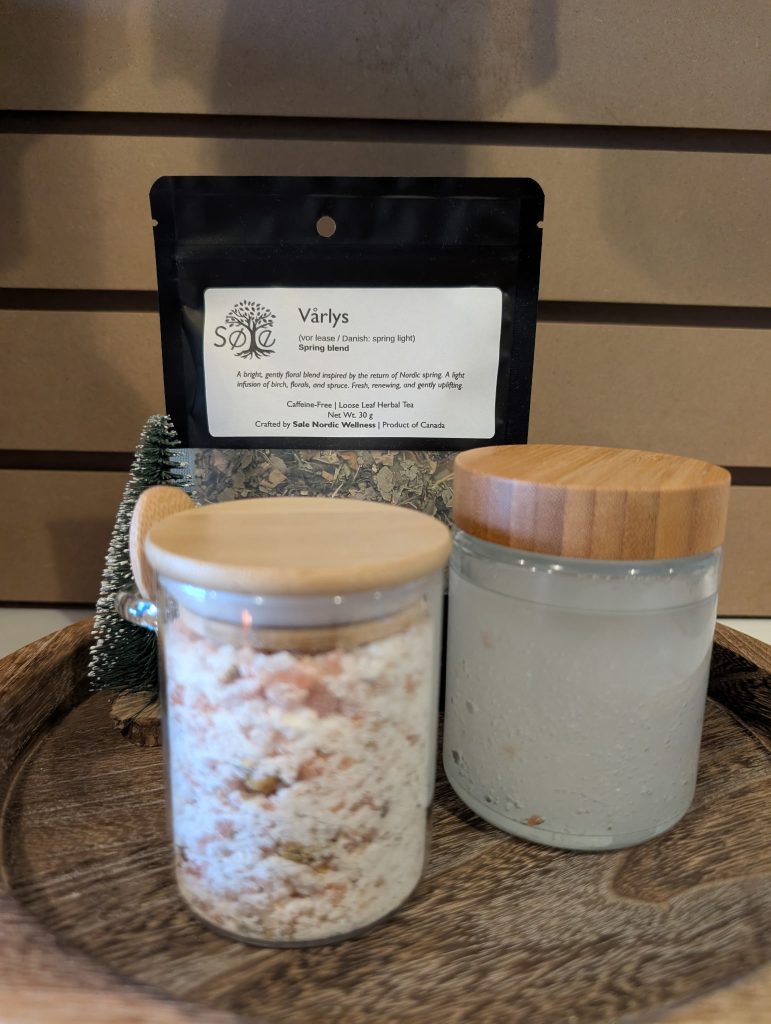 A Vårlys Tea loose tea package, a Nordic Meadow Boreal Bath Soak jar and Stødig Foot Polish jar on a wooden tray in front of a wood slat wall.